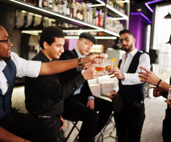Cheers! Group of handsome retro well-dressed man gangsters spend time at club. Drinking whiskey at bar counter. Multiethnic male bachelor mafia party in restaurant.
